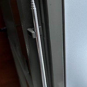 May include: A silver-coloured, multi-section telescopic aerial mounted vertically against a dark grey, textured surface. The aerial features a thin, cylindrical design with distinct segments that allow for extension, secured by two small, dark grey plastic clips.
