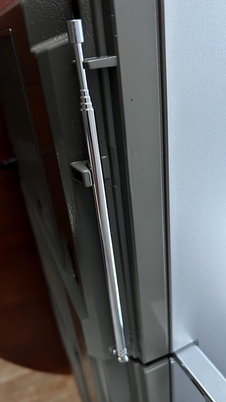 May include: A silver-coloured, multi-section telescopic aerial mounted vertically against a dark grey, textured surface. The aerial features a thin, cylindrical design with distinct segments that allow for extension, secured by two small, dark grey plastic clips.