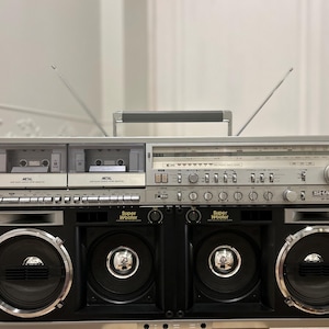 May include: A vintage silver and black Sharp GF-9000 boombox with two cassette decks, a radio tuner, and large speakers. The boombox has a handle and two antennas. The words "Super Woofer" are visible on the speaker.