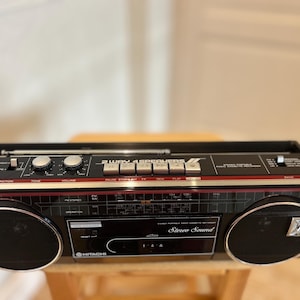 Może przedstawiać: Czarny przenośny radiomagnetofon Hitachi z 2-drożnym systemem 4 głośników, srebrną anteną teleskopową, dwoma okrągłymi głośnikami ze srebrnym wykończeniem, centralną kieszenią na kasetę oraz różnymi pokrętłami i przyciskami sterującymi.