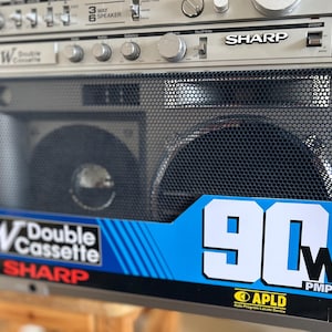 Pu&ograve; includere: Un boombox Sharp GF-767 color argento con doppio lettore di cassette, una griglia per altoparlanti in metallo nero e un adesivo blu sul davanti che riporta il testo 90W PMPO e APLD Auto Program Locate Device. Include varie manopole di controllo.