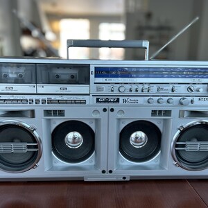 May include: A silver Sharp GF-767 boombox featuring a double cassette deck, a radio tuner with a blue display, and four front-facing speakers. The portable audio system includes a top carrying handle, a retractable antenna, and various control knobs.