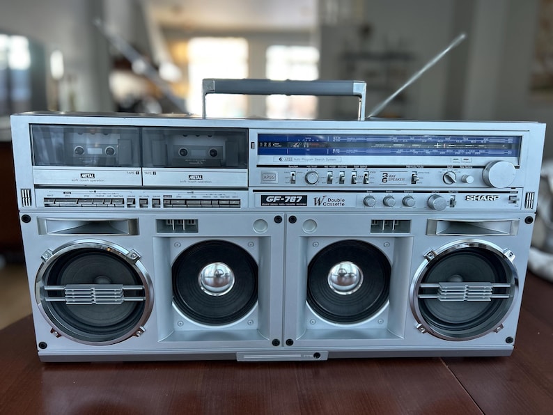 May include: A silver Sharp GF-767 boombox featuring a double cassette deck, a radio tuner with a blue display, and four front-facing speakers. The portable audio system includes a top carrying handle, a retractable antenna, and various control knobs.