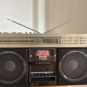 May include: A silver and black Sharp boombox featuring two large circular speakers, a central cassette deck with an APLD Sharp label, a digital display showing 1341, various control buttons, and two extendable metal antennas pointing upwards.