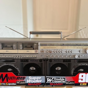 Puede incluir: Un radiocasete Sharp plateado con doble pletina, sintonizador de radio AM/FM y dos antenas extensibles. La parte frontal muestra rejillas de altavoz negras con etiquetas rojas y blancas que dicen Multi-Amp, Super Woofer y 90W PMPO.