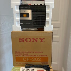 May include: A black Sony CF-302 FM/AM radio cassette recorder secured in white polystyrene packaging, resting on its original cardboard box. The box features red Sony branding and model text. Included are a black power cord and a printed instruction manual.