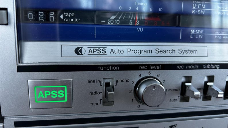 May include: A silver electronic audio device control panel featuring a bright green illuminated APSS button, a function selector switch for line in, radio, and tape, a circular rec level knob, and a tape counter display with a VU meter scale.