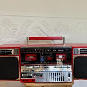 May include: A bright red portable stereo boombox featuring dual cassette decks labeled TAPE 1 and TAPE 2, a central control panel with silver buttons, a digital display, and two large black speakers on the sides with a red carrying handle on top.