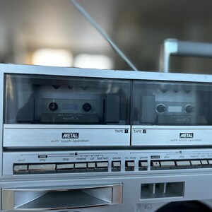 May include: A silver portable cassette player featuring dual tape decks labelled Tape 1 and Tape 2. Each deck includes a clear window, a metal soft-touch operation label, and a row of control buttons for functions like play, stop, rewind, and fast forward.