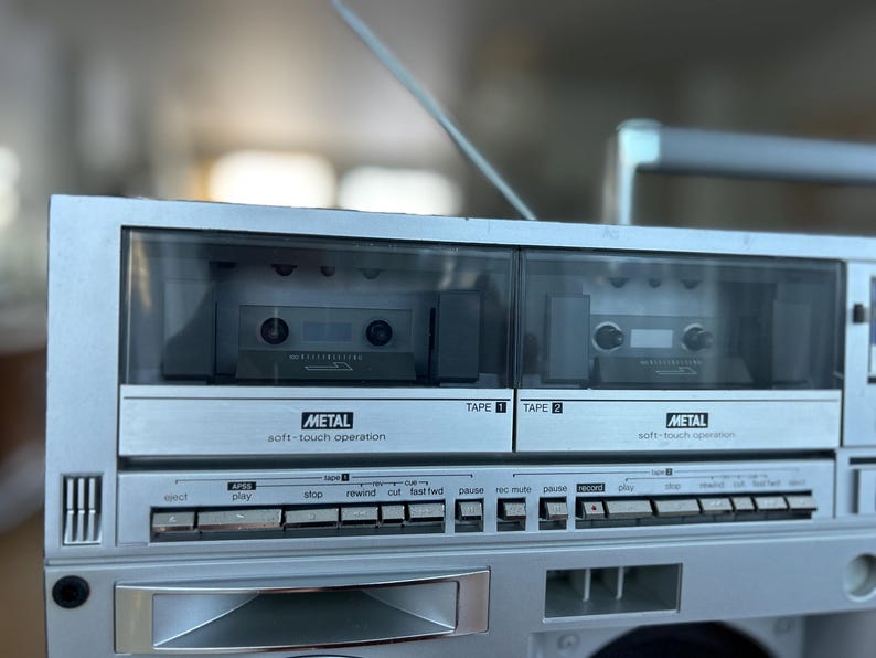 May include: A silver portable cassette player featuring dual tape decks labelled Tape 1 and Tape 2. Each deck includes a clear window, a metal soft-touch operation label, and a row of control buttons for functions like play, stop, rewind, and fast forward.