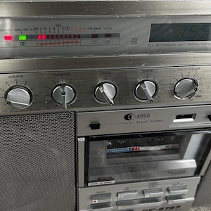 May include: Silver vintage boombox with a cassette player, radio, and various control knobs. Features a digital display and a mesh speaker cover. The boombox has the text "APSS Auto Program Search System" and "GF-9797".