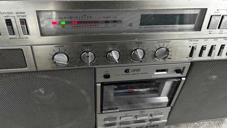 May include: Silver vintage boombox with a cassette player, radio, and various control knobs. Features a digital display and a mesh speaker cover. The boombox has the text "APSS Auto Program Search System" and "GF-9797".