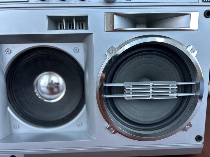 Pu&ograve; includere: Un boombox argentato con due altoparlanti circolari prominenti. L'altoparlante sinistro ha un centro metallico liscio, mentre quello destro &egrave; incorniciato da un anello cromato e presenta una griglia orizzontale argentata sul centro del cono.