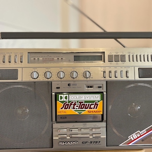 Könnte beinhalten: Ein silberner Sharp GF-9797 Ghettoblaster mit einem Kassettendeck, das ein Dolby System Soft-Touch-Etikett trägt, zwei großen runden Lautsprechern, einem Tragegriff an der Oberseite sowie verschiedenen Drehreglern und Schaltern.