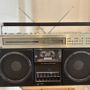 May include: A vintage silver and black Sharp GF-8000 boombox with two antennas, a cassette player, and two large speakers. The boombox has a digital clock display and a carrying handle. The text "SHARP" is visible on the front.