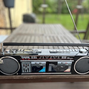 May include: A black cassette player with a silver face and a chrome handle. The player has two cassette decks, a radio tuner, and a telescopic antenna. The player is sitting on a wooden table.