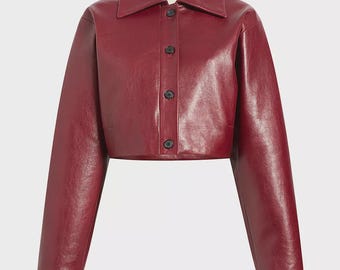May include: A cropped, burgundy-colored jacket with a collared neckline and long sleeves. The jacket features a button-up front with black buttons. The material has a slight sheen, suggesting a leather or faux-leather finish. The jacket is a stylish outerwear piece.