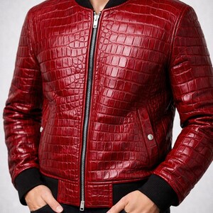 Puede incluir: Chaqueta bomber de cuero rojo con textura de piel de cocodrilo. La chaqueta tiene cuello, puños y dobladillo de canalé negro, y cierre de cremallera plateada. La chaqueta tiene dos bolsillos laterales y se usa con pantalones negros.