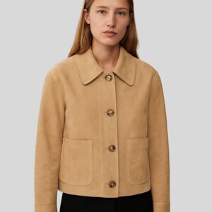 May include: A tan suede jacket with a collared neckline, button-up front, and two front pockets. The jacket has long sleeves with buttoned cuffs. The model is wearing a black top.