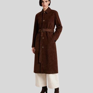 May include: A long-length dark brown suede coat featuring a classic collar, a front button closure, and a matching suede belt with a buckle at the waist. The coat is paired with wide-leg off-white trousers and dark brown ankle boots.