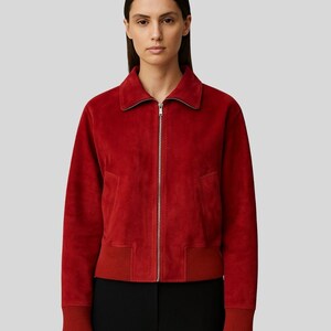 May include: A red suede bomber jacket with a silver zipper closure. The jacket has a collar, side pockets, and ribbed cuffs and hem. The sleeves are long, and the jacket is paired with black pants.