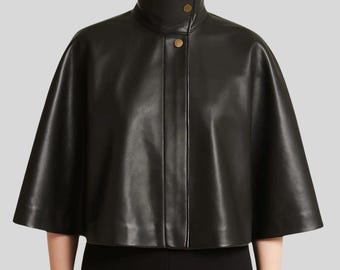 May include: A black leather cape with a high collar and two gold-colored buttons. The cape has a short, wide silhouette and is made of a smooth, shiny material. The garment is a stylish outerwear piece.