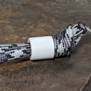 May include: A white cylindrical object is positioned on a black, white, and grey patterned rope, which is knotted at the end. The background is a dark, textured surface.