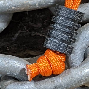 Puede incluir: Primer plano de un cierre de cord&oacute;n negro y gris con un cord&oacute;n naranja brillante enhebrado. El cierre de cord&oacute;n est&aacute; unido a una cadena gruesa de plata.