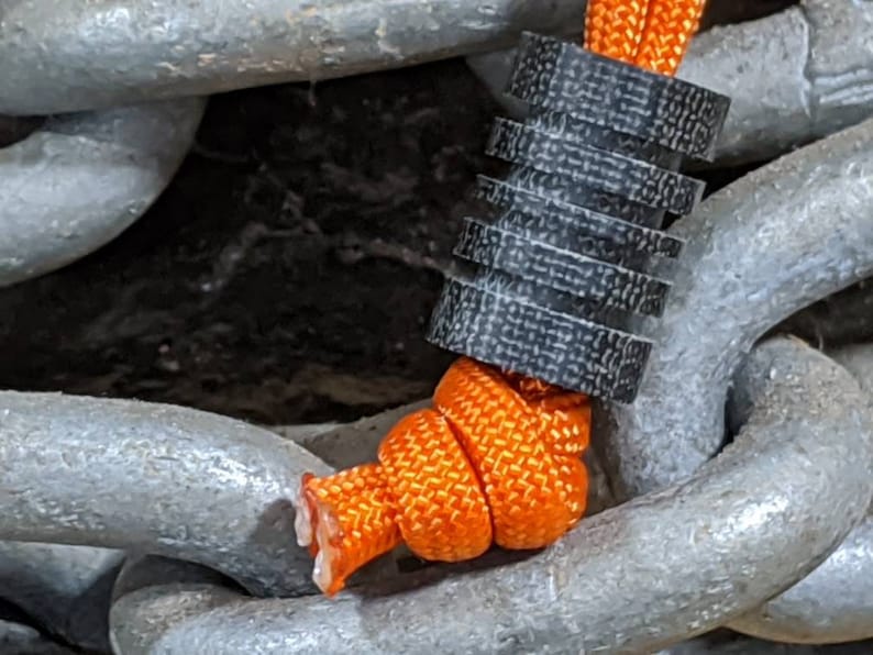 Puede incluir: Primer plano de un cierre de cord&oacute;n negro y gris con un cord&oacute;n naranja brillante enhebrado. El cierre de cord&oacute;n est&aacute; unido a una cadena gruesa de plata.