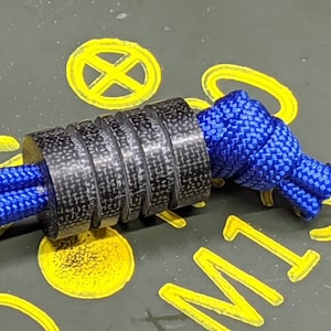 Puede incluir: Un toggle de paracord de fibra de carbono negro con un nudo de paracord azul. El toggle es cil&iacute;ndrico y tiene una superficie texturizada.