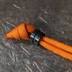 May include: A close-up of an orange paracord tied with a knot and secured with a black plastic toggle.