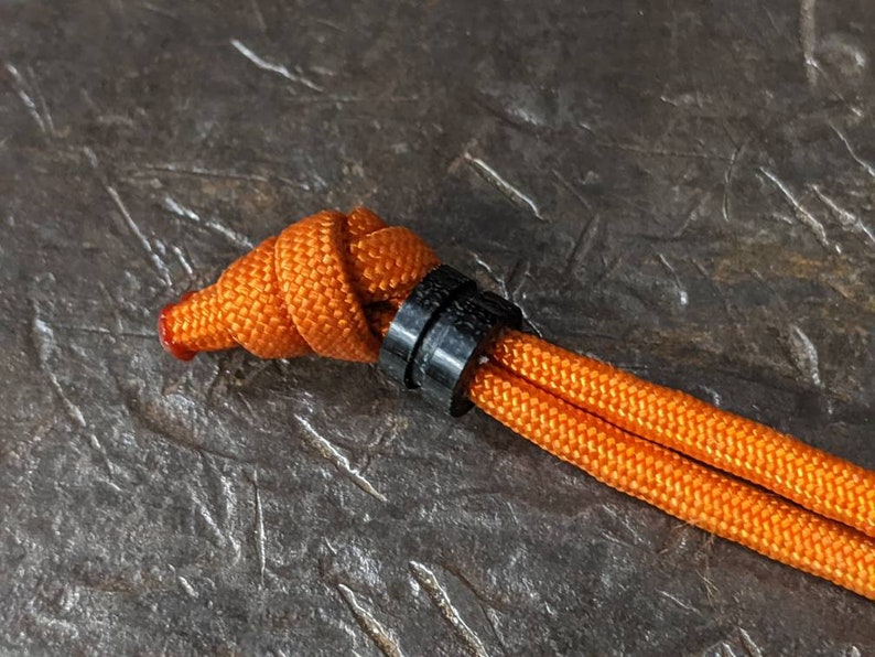 May include: A close-up of an orange paracord tied with a knot and secured with a black plastic toggle.