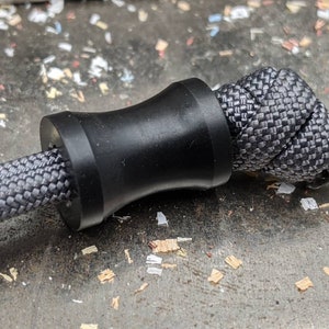 May include: A black paracord bracelet with a black bead. The bead is a cylinder with a slightly wider middle section. The paracord is woven in a tight, intricate pattern.