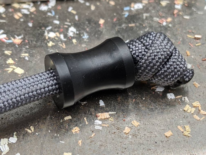 May include: A black paracord bracelet with a black bead. The bead is a cylinder with a slightly wider middle section. The paracord is woven in a tight, intricate pattern.