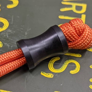 May include: A black cord end stop with a red paracord threaded through it. The cord end stop is a cylindrical shape with a smooth, black finish.
