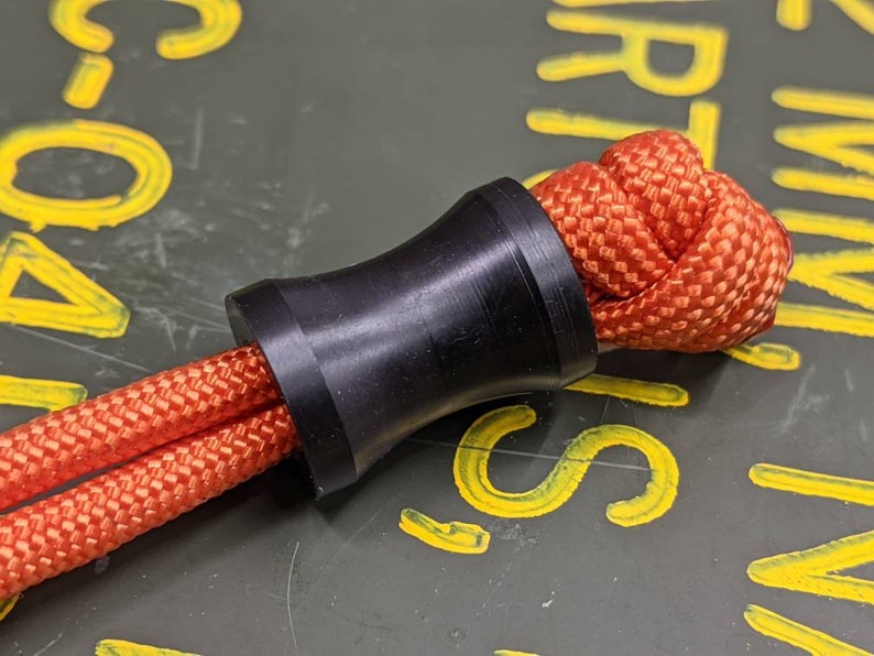 May include: A black cord end stop with a red paracord threaded through it. The cord end stop is a cylindrical shape with a smooth, black finish.
