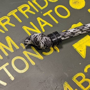 May include: A black and white paracord with a black plastic toggle. The paracord is lying on a green surface with yellow markings that say "CARTRIDGE MM NATO" and "AB33".