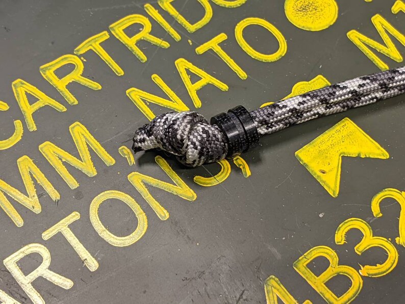 May include: A black and white paracord with a black plastic toggle. The paracord is lying on a green surface with yellow markings that say "CARTRIDGE MM NATO" and "AB33".