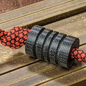 Puede incluir: Una cuenta de fibra de carbono negra y gris con una cuerda enhebrada a trav&eacute;s de ella. La cuerda es roja y negra con un patr&oacute;n de cuadros.