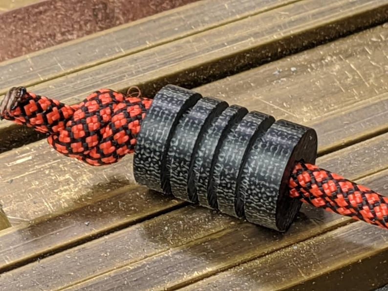 Puede incluir: Una cuenta de fibra de carbono negra y gris con una cuerda enhebrada a trav&eacute;s de ella. La cuerda es roja y negra con un patr&oacute;n de cuadros.