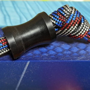 May include: A black plastic cord stop with a braided rope threaded through it. The rope is red, white, and blue.