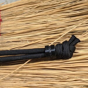 May include: Close-up of a black paracord with a black plastic bead and a knot tied in the cord. The cord is resting on a brown, woven surface.