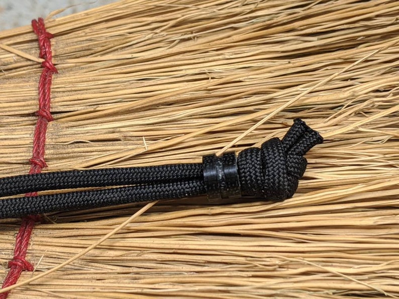 May include: Close-up of a black paracord with a black plastic bead and a knot tied in the cord. The cord is resting on a brown, woven surface.