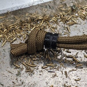 May include: A brown paracord cord with a black cord slider. The cord is tied in a knot and is lying on a surface covered in gold metal shavings.