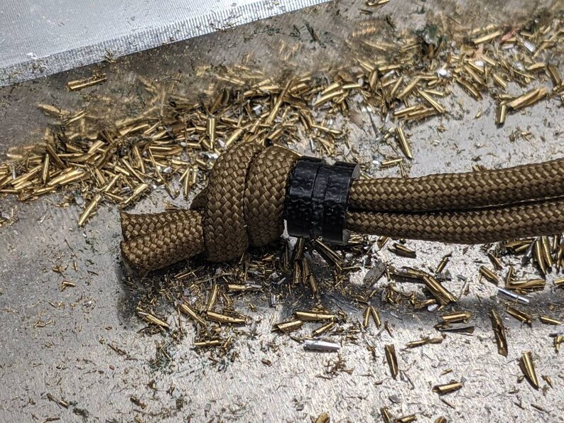 May include: A brown paracord cord with a black cord slider. The cord is tied in a knot and is lying on a surface covered in gold metal shavings.