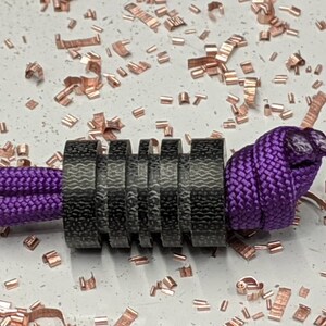 Puede incluir: Un brazalete de paracord negro y morado con una cuenta de metal negro. La cuenta est&aacute; hecha de varios anillos apilados.