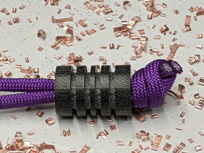 Puede incluir: Un brazalete de paracord negro y morado con una cuenta de metal negro. La cuenta est&aacute; hecha de varios anillos apilados.
