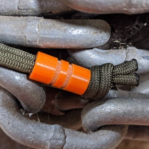 May include: A close-up of an orange paracord bead with a green paracord rope attached to it. The bead is made of plastic and has a textured surface. The rope is wrapped around a metal chain.