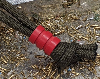 May include: A red plastic cord lock with a black paracord rope threaded through it. The cord lock is on a surface covered in small pieces of metal.