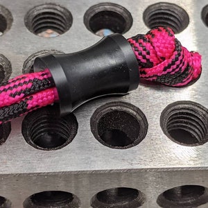May include: A black cord stopper with a pink and black braided cord threaded through it. The cord stopper is sitting on a metal surface with multiple holes.